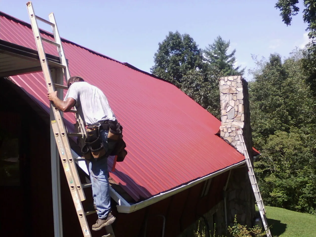 Roofer on a ladder beside a red metal roof and stone chimney — Soffit & Fascia Repair by Roofing Brothers in Rebersburg